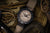 Herrenarmbanduhr mit beigem Textilarmband, schwarzer Lünette und beigem Zifferblatt.