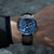 Alt text: Orologio Naos con quadrante a fasi lunari, cassa in acciaio inossidabile e cinturino in pelle marrone.
