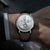 Alt text: Orologio Naos con quadrante a fasi lunari, cassa in acciaio inossidabile e cinturino in pelle marrone.