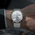 Alt text: Orologio Naos con quadrante a fasi lunari, cassa in acciaio inossidabile e cinturino in pelle marrone.