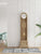 Orologio a pendolo in legno chiaro con quadrante rotondo, anta in vetro e pomello bianco