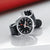 Mondaine Stop2Go-Uhr schräg aufgestellt: schwarzes Zifferblatt, silbernes Gehäuse, schwarzes Lederarmband