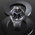 Orologio da uomo frontale con quadrante nero, cassa argentata e cinturino in metallo.