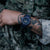 Orologio da uomo al polso con abbigliamento mimetico: quadrante blu, cinturino scuro, indici luminosi visibili.