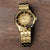 Herrenarmbanduhr mit goldfarbenem Edelstahlband, rundem goldenem Zifferblatt und Knopf an der Seite.