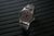 Herrenarmbanduhr mit leuchtenden Markierungen auf dem Zifferblatt und dunklem Lederarmband.